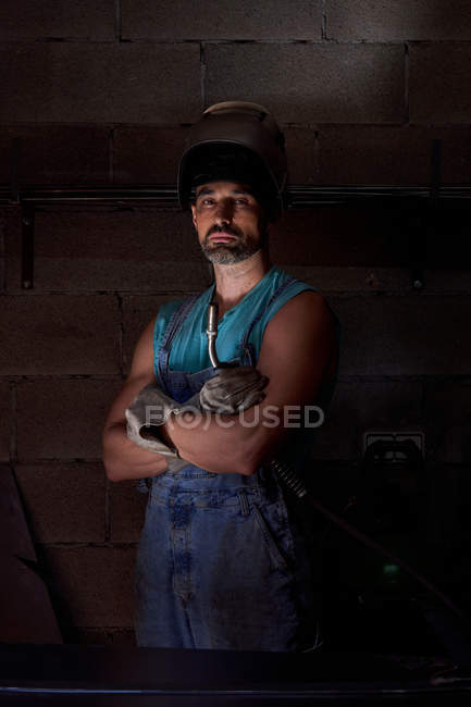 Serious confident bearded workman in welding helmet on head wearing overalls and protective gloves standing with arms crossed with dark stone wall in background — Stock Photo