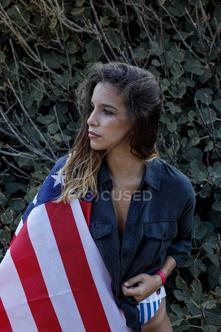 Alegre encaracolado fêmea hipster com bandeira americana olhando para longe por plantas verdes — Fotografia de Stock