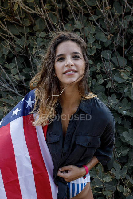 Cheerful curly-haired female hipster with American flag looking in camera by green plants — Stock Photo