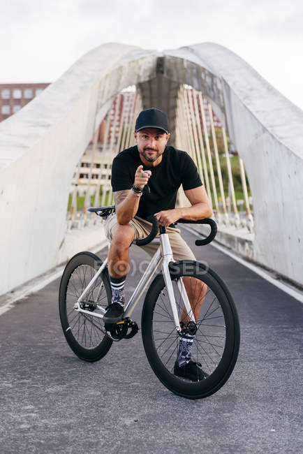 Happy adult bearded man in black cap wearing black shirt and beige shorts sitting resting on bicycle across footbridge in city looking at camera — Stock Photo