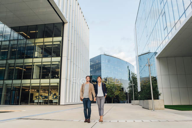 Знизу щаслива пара колег, тримаючись за руки і йдучи разом за межами сучасної будівлі на міській вулиці після роботи — стокове фото