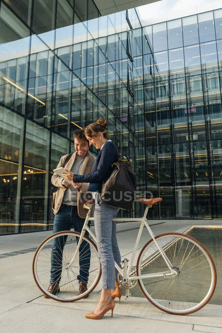 Cheerful man and woman with bicycle smiling and looking at a tablet while communicating outside office building — Stock Photo