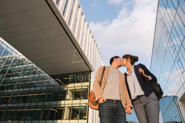 Von unten glückliche Paar-Kollegen, die sich küssen, während sie nach Feierabend vor einem modernen Gebäude an der Stadtstraße stehen — Stockfoto