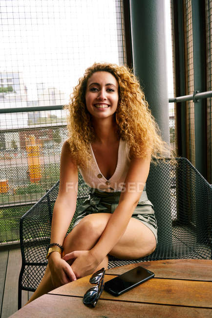 Cheerful curly woman comfortably sitting in metal chair with sunglasses and smartphone at table and looking at camera in airport of Texas — Stock Photo