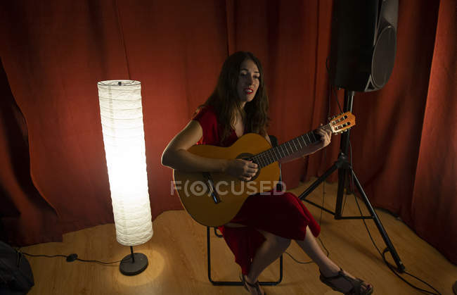 Encantadora mujer artística en vestido rojo tocando la canción en la guitarra en el escenario con luz cálida - foto de stock