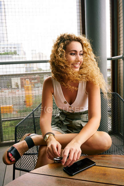 Cheerful curly woman comfortably sitting in metal chair with sunglasses and smartphone at table and looking away in airport of Texas — Stock Photo