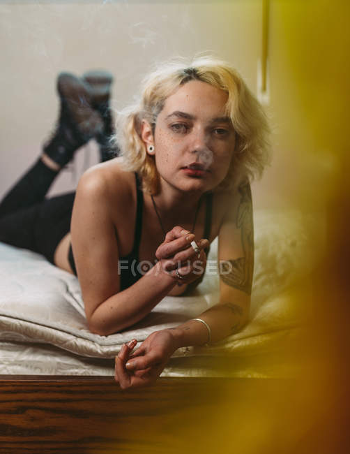 Mujer en negro ropa casual y zapatos fumar cigarrillo y reflexionar mientras está acostado solo en la cama y mirando a la cámara - foto de stock