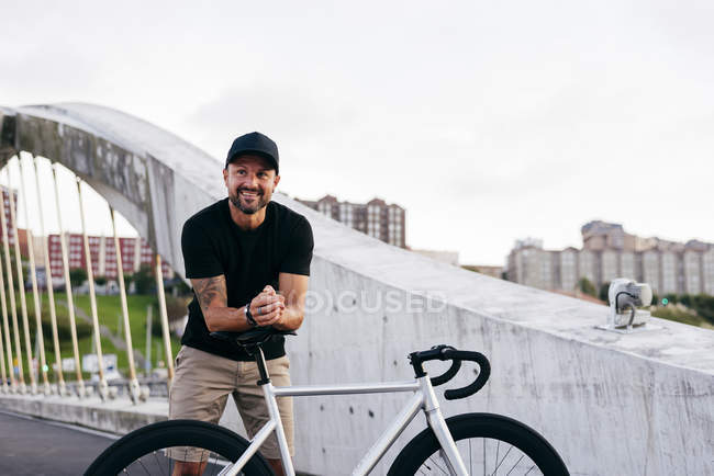 Homem barbudo adulto feliz em boné preto vestindo camisa preta e shorts bege de pé inclinado na bicicleta sentar-se em toda a ponte na cidade olhando para longe — Fotografia de Stock