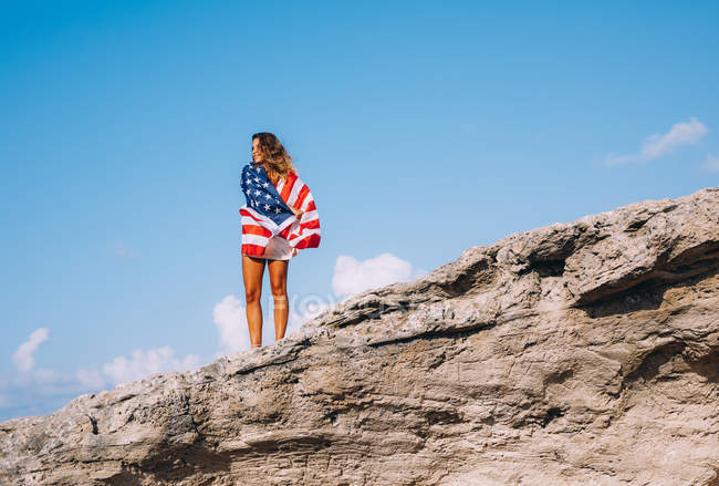 Cheerful woman wrapped in American flag standing on rocky cliffs against blue sky — Stock Photo