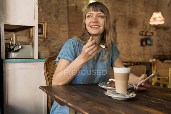 Блондинка весела щаслива жінка з чубчиком тримає ложку з шматочком торта і сидить за столом з кавою і десертом — стокове фото