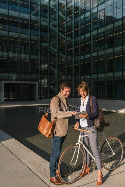 Cheerful man and woman with bicycle smiling and looking at a tablet while communicating outside office building in city street — Stock Photo
