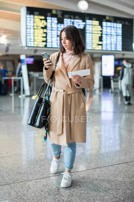 Випадкова молода жінка в стильному пальто стоїть з ручною поклажею і квитком під час смс на мобільний телефон в аеропорту — стокове фото
