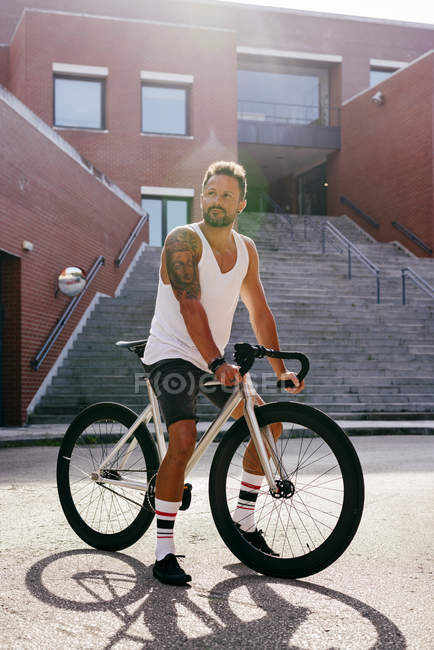 Sportive man wearing white sleeveless shirt and black shorts sitting on bicycle between buildings near stairs — Stock Photo