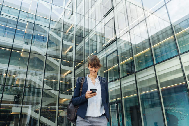 Низький кут щасливої бізнес-леді, використовуючи смартфон, стоячи за межами бізнес-центру на міській вулиці — стокове фото