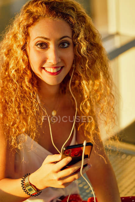 Piacevole donna allegra in auricolare ascoltare musica con il telefono cellulare mentre si raffredda sulla panchina metallica in aeroporto del Texas — Foto stock
