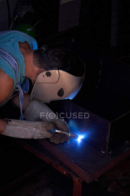 Side view of craftsman in protective face mask wearing overalls and gloves working in workshop and soldering metal construction — Stock Photo