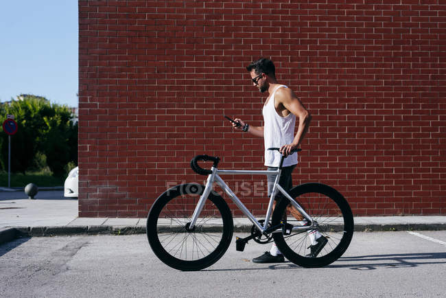 Vista lateral do belo ciclista masculino em sportswear e óculos de sol usando smartphone enquanto está de pé com bicicleta ao lado da parede de tijolo vermelho — Fotografia de Stock