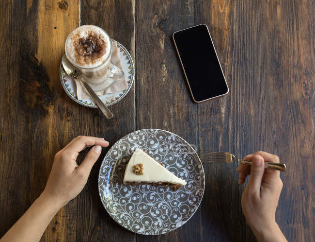Female holding fork as sitting at table with blank screen smartphone next to coffee and cake on plate with ornament — Stock Photo