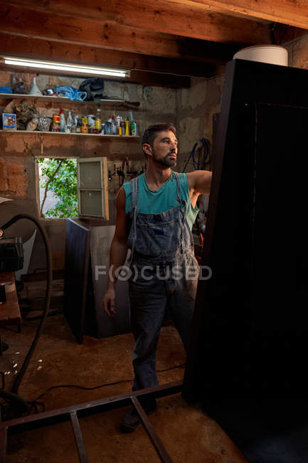 Hombre experto de mediana edad en overoles de mezclilla trabajando con muebles en el taller - foto de stock