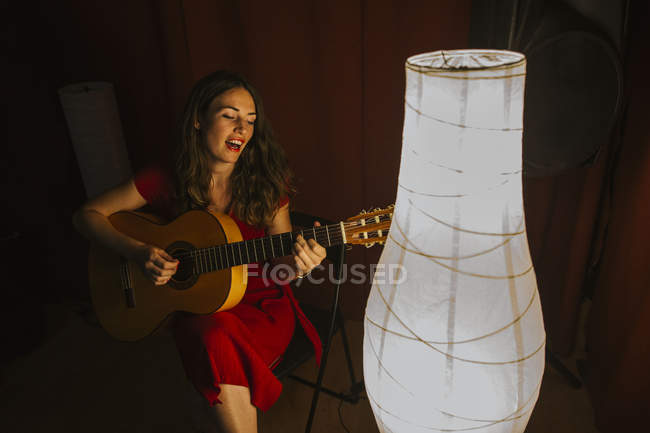 Зверху талановита жінка в червоній сукні виконує пісню і грає на гітарі в теплій освітленій сцені поблизу білої лампи — стокове фото