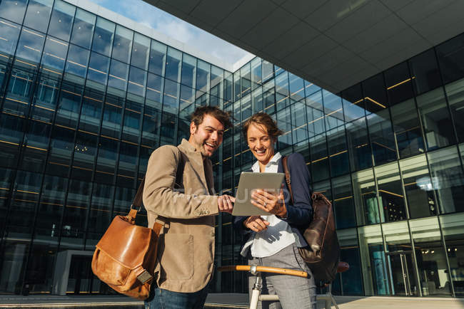 Empresarios optimistas sonriendo y navegando por las redes sociales en la tableta mientras están de pie fuera del edificio moderno - foto de stock