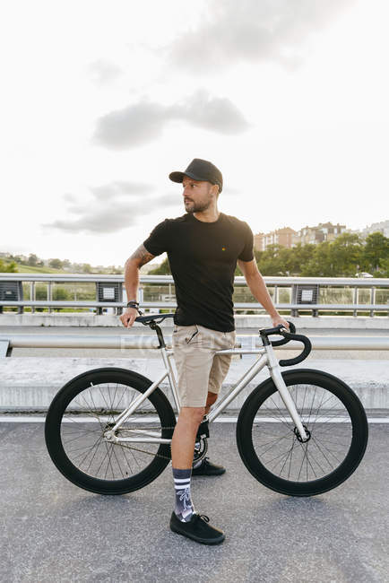 Счастливый взрослый бородатый мужчина в черной кепке в черной рубашке и бежевых шортах отдыхает с велосипедом через пешеходный мост в городе глядя в сторону — стоковое фото