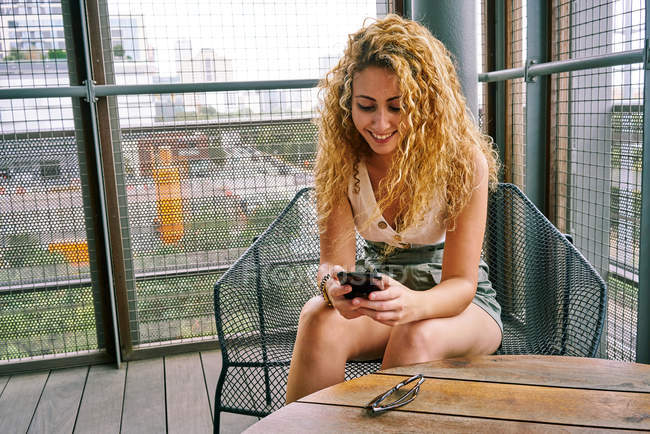 Sorrindo mulher encaracolado refrigeração na moderna área de espera sentado em cadeira de metal e surf telefone celular Texas — Fotografia de Stock