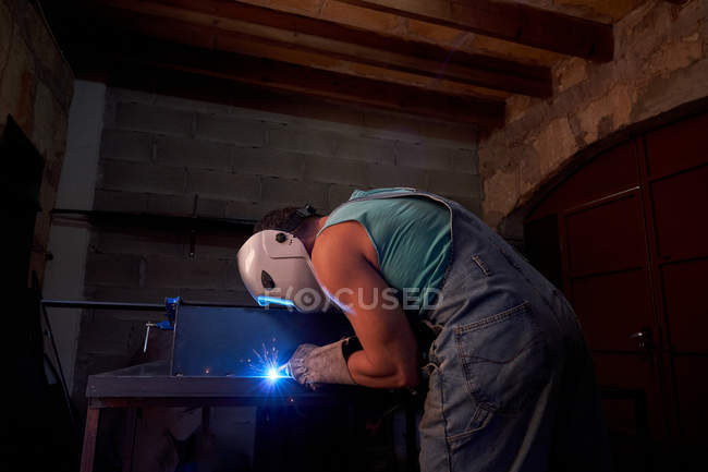 Vue latérale de l'artisan en masque protecteur portant des combinaisons et des gants travaillant en atelier et soudant la construction métallique — Photo de stock