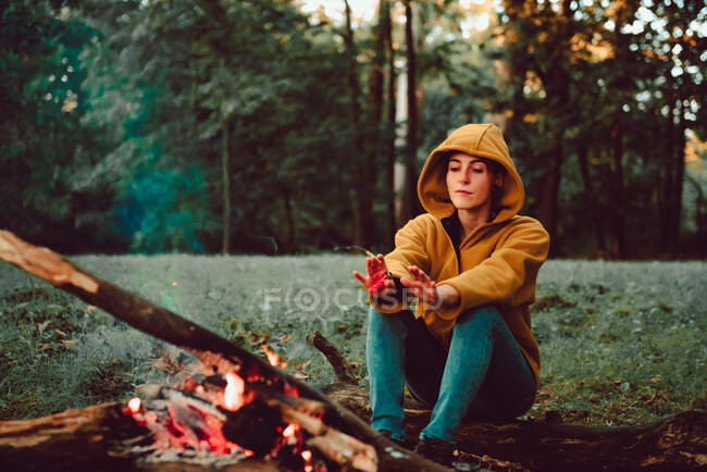 Pensive relaxed adult female in hoodie focusing on flame and contemplating while sitting alone and warming near bonfire on glade with lush green grass in tranquil dense forest — Stock Photo