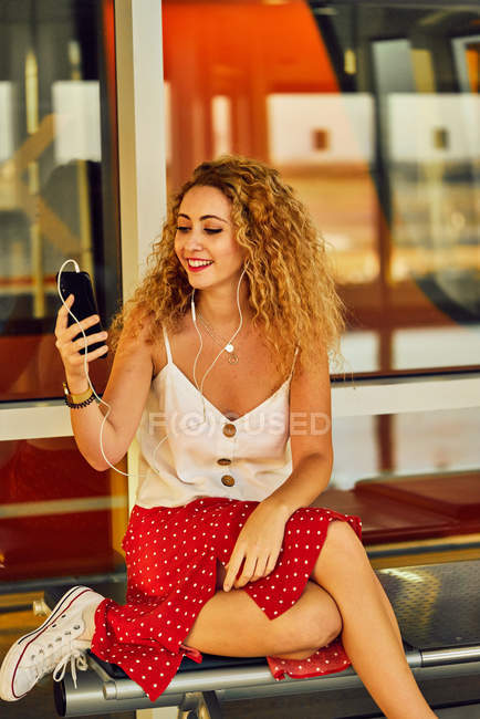 Agradable mujer alegre en auriculares escuchando música con teléfono móvil mientras se enfría en un banco de metal en el aeropuerto de Texas - foto de stock
