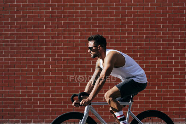 Сучасний чоловічий велосипедист у спортивній манері та сонцезахисних окулярах їде на велосипеді біля червоної цегляної стіни. — стокове фото