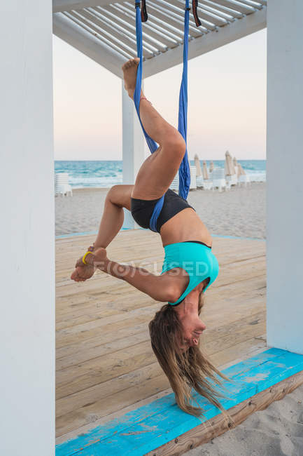 Sportive power woman hanging upside down on hammock for aerial yoga holding on leg in wooden stand on beach — Stock Photo