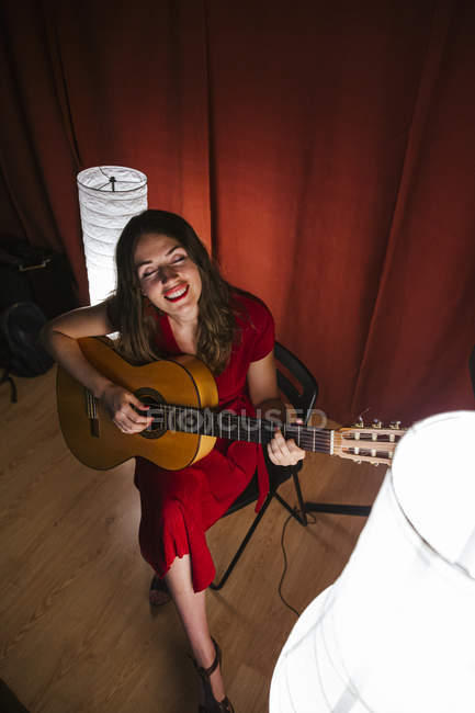 De cima talentosa mulher em vestido vermelho tocando música e guitarra no palco iluminado quente lâmpada branca nas proximidades — Fotografia de Stock