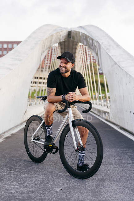 Hombre barbudo adulto feliz en gorra negra con camisa negra y pantalones cortos beige sentado descansando en bicicleta cruzando la pasarela en la ciudad mirando hacia otro lado - foto de stock