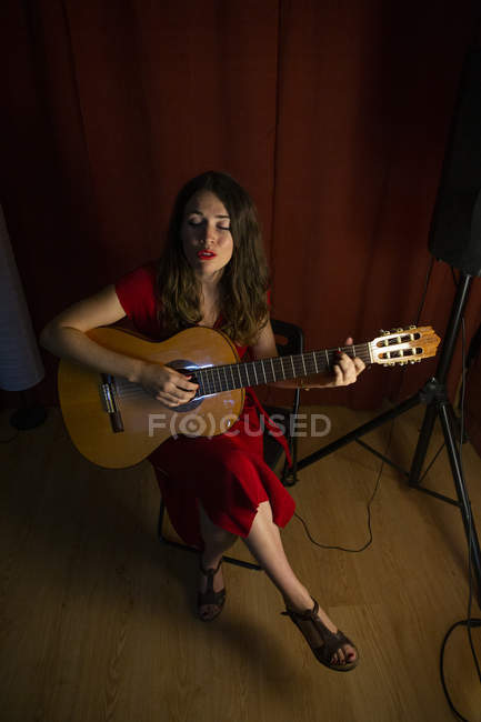 D'en haut femme talentueuse en robe rouge chantant et jouant de la guitare sur scène éclairée chaude lampe blanche à proximité — Photo de stock