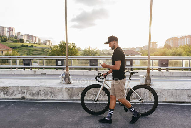 Side view of young casual bearded cyclist in black cap using phone while walking with bike on road with city buildings on background — Stock Photo