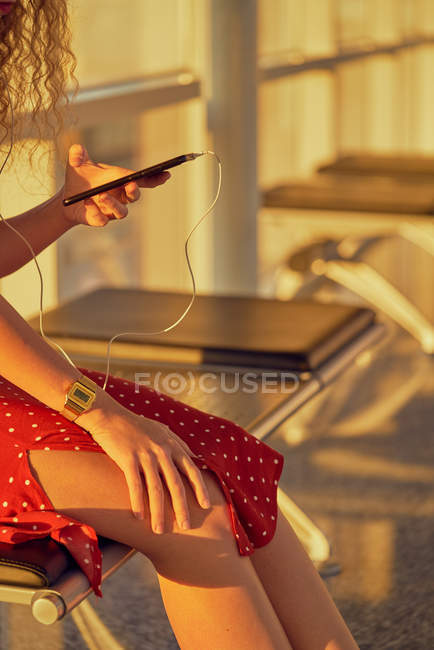 Зображення жінки в навушниках, яка слухає музику за допомогою мобільного телефону, коли холоне на металевій лавці в аеропорту Техасу. — стокове фото