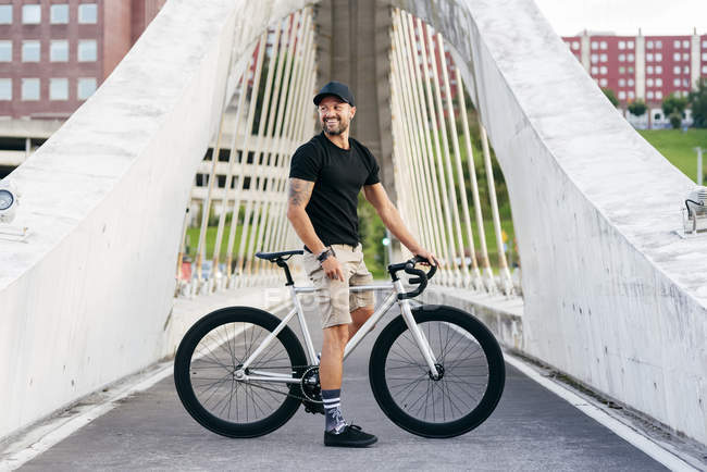 Happy adult bearded man in black cap wearing black shirt and beige shorts standing with bike across footbridge in city looking away — Stock Photo