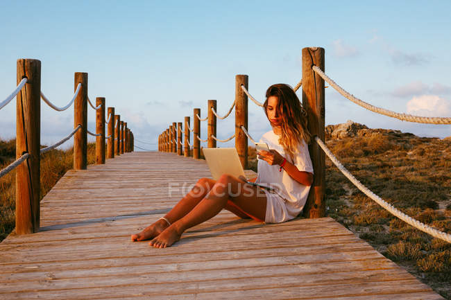 Freelancer de vacaciones en camisa blanca sentado en puente vacío y trabajando con portátil y usando teléfono móvil sobre fondo azul del cielo - foto de stock