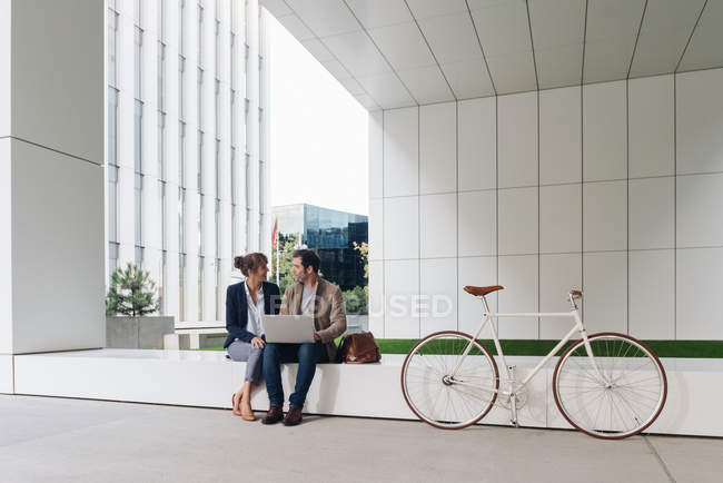 Delighted businesspeople smiling and browsing laptop together while sitting outside modern building near bicycle on city street — Stock Photo