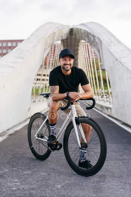 Счастливый взрослый бородатый мужчина в черной кепке в черной рубашке и бежевых шортах сидит на велосипеде через пешеходный мост в городе и смотрит в сторону — стоковое фото