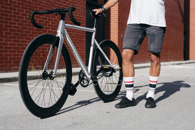 Image recadrée de cycliste en vêtements de sport à l'aide d'un smartphone tout en restant debout avec le vélo à côté du mur de briques rouges — Photo de stock