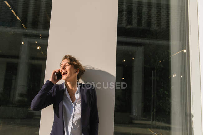 Optimistic businesswoman in dark jacket smiling and looking away while leaning on building wall on city street and answering phone call — Stock Photo
