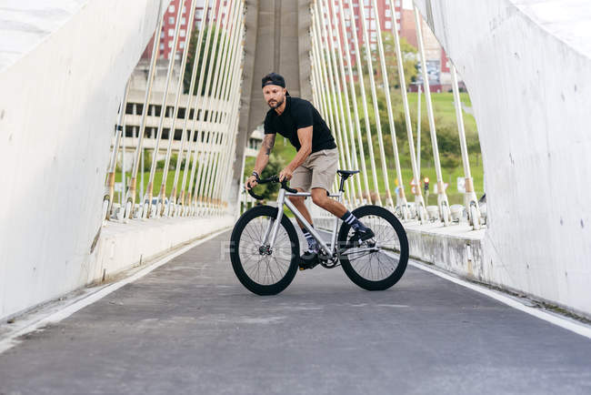 Uomo barbuto adulto in berretto nero con camicia nera e pantaloncini beige in sella a una bicicletta attraverso il ponte pedonale in città guardando altrove — Foto stock