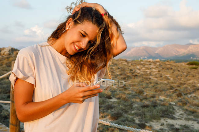Voyageur en chemise blanche décontractée toucher les cheveux tout en surfant sur le web sur téléphone mobile avec beau paysage sur fond — Photo de stock