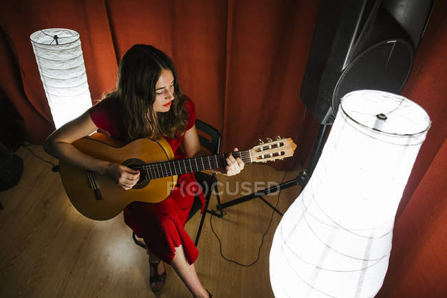 Desde arriba mujer talentosa en vestido rojo interpretando canción y tocando la guitarra en el escenario iluminado cálido lámpara blanca cercana - foto de stock