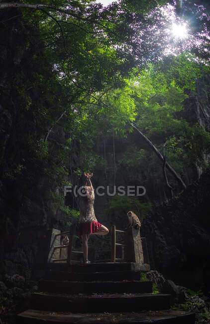Back view of unrecognizable female standing on one leg on ancient pedestal and meditating with raised arms in middle of green forest in Thailand — Stock Photo