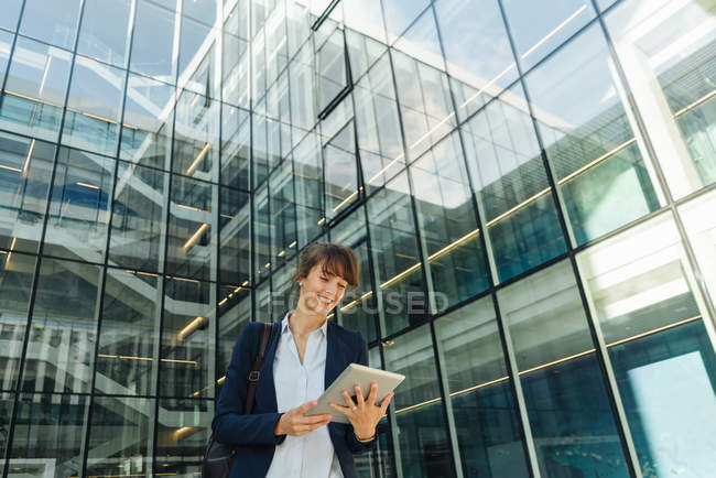 Bajo ángulo de empresario alegre sonriendo y tableta de navegación mientras está de pie fuera del edificio con paredes de vidrio en el centro de la ciudad - foto de stock