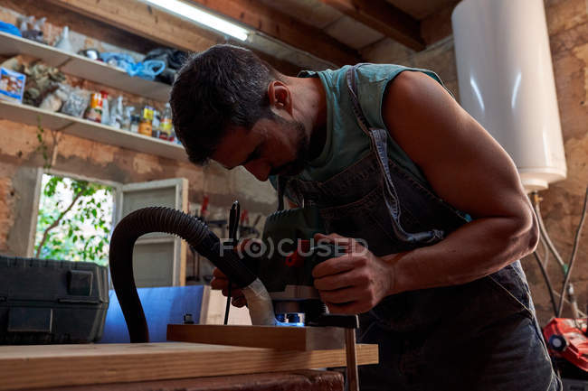 Carpintero procesando detalle de madera con fresadora mientras trabaja en taller - foto de stock