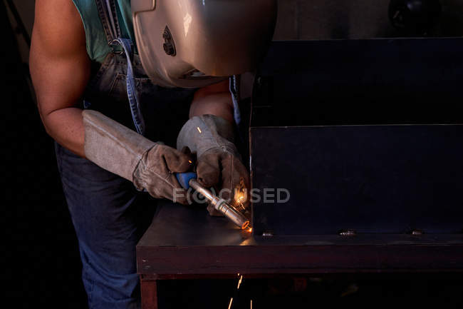 Ремесленник в защитной маске для лица в комбинезонах и перчатках, работающих в мастерской и паяльной металлической конструкции — стоковое фото